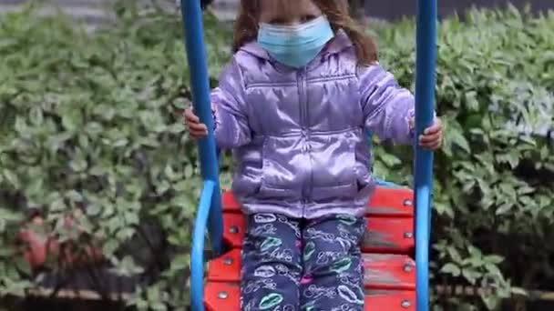 의학적 푸른 마스크를 쓴 귀여운 금발 소녀가 운동장에서 그네를 타고 있습니다. 19 개 지방 자치체를 관할 한다. 고립 된 아이들. FullHD 영상. — 비디오