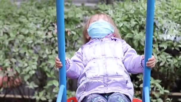 의학적 푸른 마스크를 쓴 귀여운 금발 소녀가 운동장에서 그네를 타고 있습니다. 19 개 지방 자치체를 관할 한다. 고립 된 아이들. FullHD 영상. — 비디오