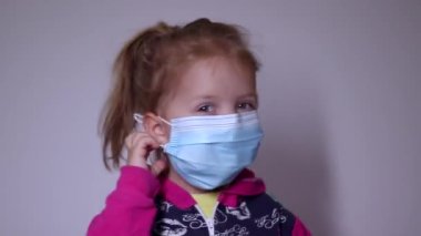 Mavi tıbbi maskeli küçük sarışın kız üzgün. Maskeyi çıkar. Pembe kazaklı. Covid 19 pandemia. Çocuklar karantinada. Tam HD görüntüler.