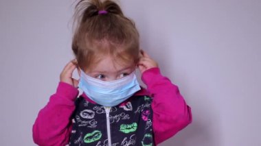 Mavi tıbbi maskeli küçük sarışın kız. Giymeye çalışıyorum. Pembe kazaklı. Covid 19 pandemia. Çocuklar karantinada. Tam HD görüntüler.