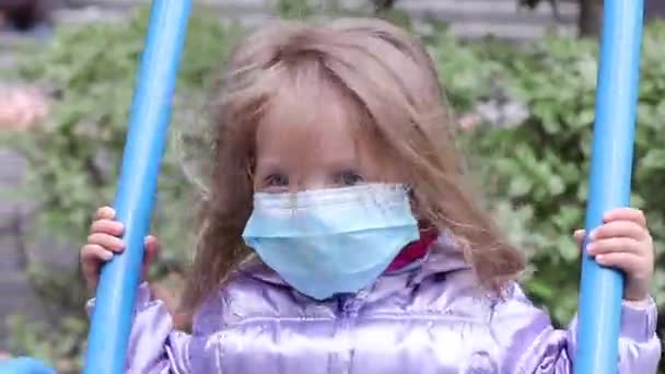 Kleine süße blonde Mädchen in einer medizinischen blauen Maske reitet eine Schaukel auf dem Spielplatz. Covid 19 Pandemie. Kinder in Isolation. FullHD-Aufnahmen. — Stockvideo