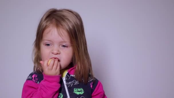 Bonita niña rubia se come una galleta sobre fondo grisáceo. Imágenes Full HD. — Vídeo de stock