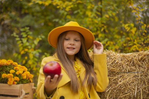 秋の飾りの横にあるオレンジの帽子 ドレス コートのかわいい十代のブルネットの女の子は赤いリンゴを持っています — ストック写真
