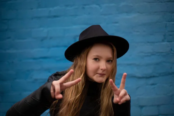 在蓝色砖墙的背景上 身穿皮革夹克 头戴黑色帽子的可爱的黑发少女的画像 显示出和平的迹象 — 图库照片