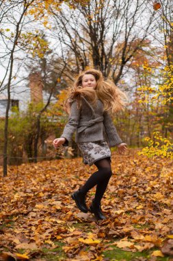 Gri montlu güzel esmer kız sonbahar ormanına atlıyor. Sıcaklık, sonbahar, eğlence.