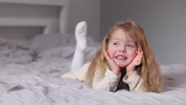 Šťastná hezká malá blondýnka v béžových šatech ležící na posteli v šedé ložnici. ranní rutina, dětství, batole, dcera. FullHD záznam — Stock video
