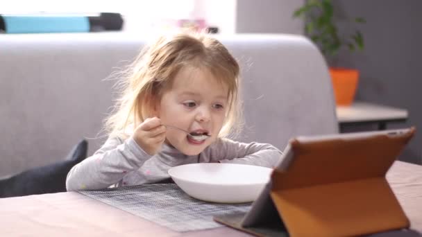 Pretty little blonde girl in pajama eating milk porridge with asterisk vermicelli, looking in the tablet. breakfast, morning, home. FullHD footage — Vídeos de Stock