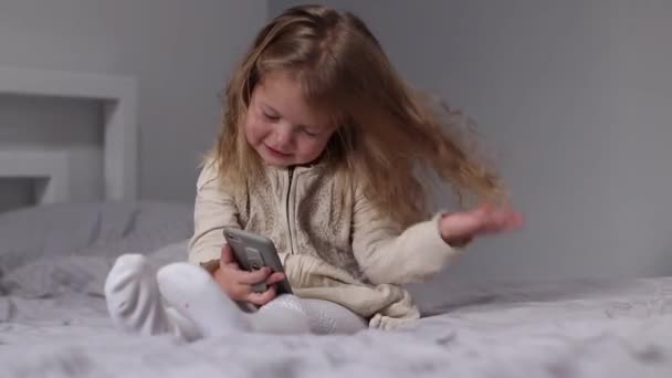 Pretty little blonde girl in dress looking in phone in gray bedroom. morning routine, childhood, toddler, daughter. FullHD footage — Vídeos de Stock