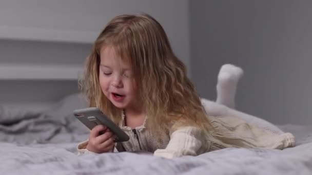 Pretty little blonde girl in dress looking in phone in gray bedroom. morning routine, childhood, toddler, daughter. FullHD footage — Vídeo de stock