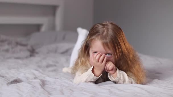 Feliz bonita niña rubia en vestido beige acostado en una cama en el dormitorio gris. rutina de la mañana, infancia, niño, hija. Imágenes de FullHD — Vídeos de Stock