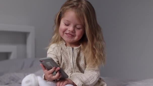 Pretty little blonde girl in dress looking in phone in gray bedroom. morning routine, childhood, toddler, daughter. FullHD footage — Vídeos de Stock