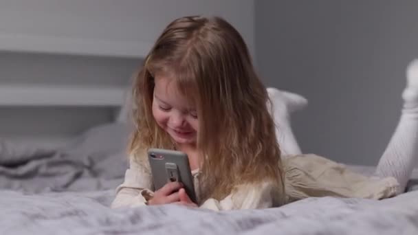 Pretty little blonde girl in dress looking in phone in gray bedroom. morning routine, childhood, toddler, daughter. FullHD footage — Vídeo de stock