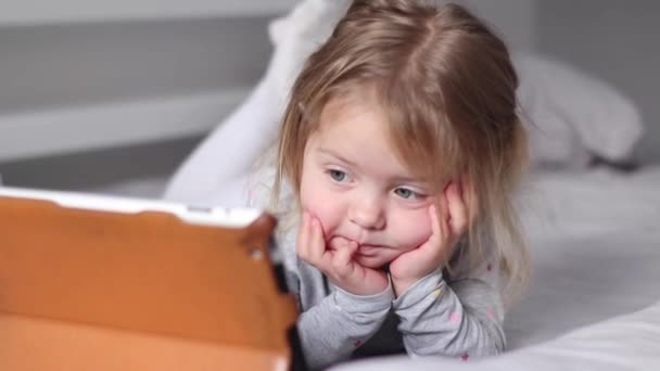 Pijamalı güzel sarışın kız gri yataktaki tablette çizgi film izliyor. Sabah rutini, çocukluk, bebek, kız. Tam HD görüntüler — Stok video