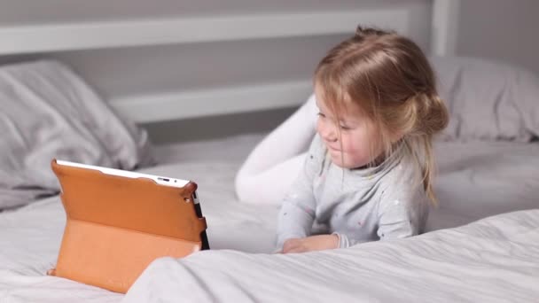 Bonita niña rubia en pijama viendo dibujos animados en la tableta en la cama gris. rutina de la mañana, infancia, niño, hija. Imágenes de FullHD — Vídeos de Stock