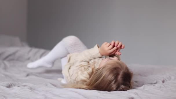 Feliz bonita niña rubia en vestido beige acostado en una cama en el dormitorio gris. rutina de la mañana, infancia, niño, hija. Imágenes de FullHD — Vídeos de Stock