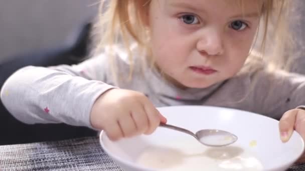 Pretty little blonde girl in pajama eating milk porridge with asterisk vermicelli. breakfast, morning, home. FullHD footage — Stockvideo