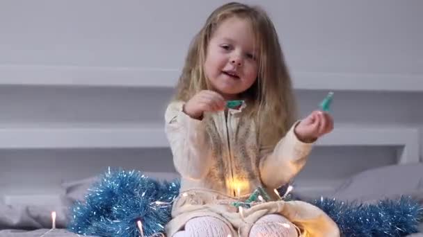 Bonita niña jugando con pequeños árboles de Navidad y guirnalda acostada en la cama en el dormitorio gris. Año Nuevo, infancia, hija. Imágenes de FullHD — Vídeos de Stock