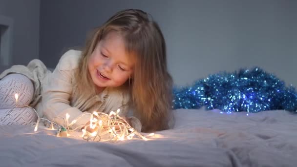 Bonita niña jugando con pequeños árboles de Navidad y guirnalda acostada en la cama en el dormitorio gris. Año Nuevo, infancia, hija. Imágenes de FullHD — Vídeos de Stock
