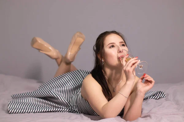 灰色の寝室のベッドの上にシャンパンのガラスと夜のカクテルドレスで幸せな魅力的なブルネットの女性 アフターパーティー孤独 コピースペース — ストック写真