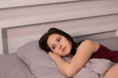 Üzgün, uykulu esmer kız, bordo bluzlu, gri çarşafların altında, yatakta. Sabah rutini. Uyurken kötü bir rüya gördüm. Boşluğu kopyala