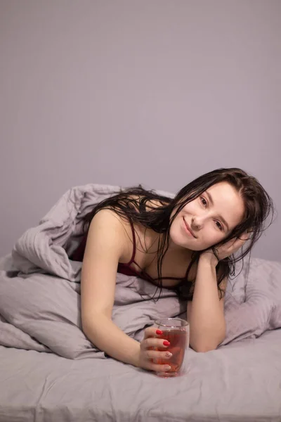 魅力的な眠いブルネットの女性グレーのベッドリネンとベッドの中でお茶のグラスカップ 朝のルーティン コピースペース — ストック写真