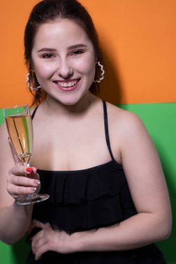 Siyah elbiseli çekici esmer kadın elinde bir kadeh şampanya ve parlak yeşil turuncu arka planda bir nişan yüzüğü var. Düğün konsepti. Nişan. Soruyu cevaplıyorum. gelin