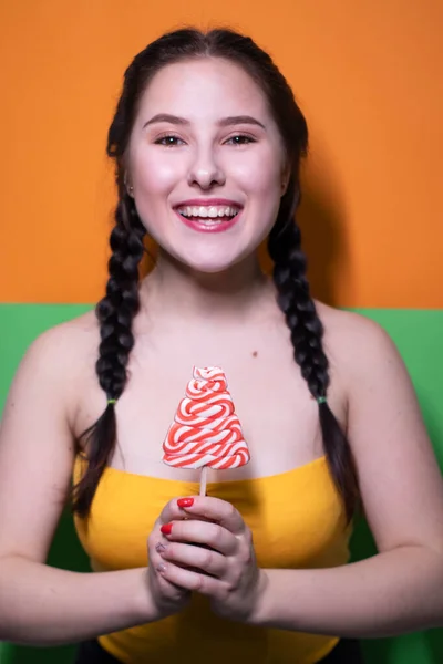 鮮やかな緑とオレンジの背景に赤い大きなロリポップを保持黄色の上に魅力的なブルネットの女性 お菓子のコンセプト キャンディ コピースペース — ストック写真