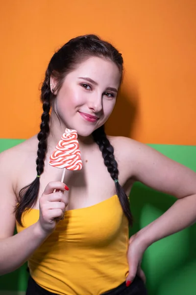 鮮やかな緑とオレンジの背景に赤い大きなロリポップを保持黄色の上に魅力的なブルネットの女性 お菓子のコンセプト キャンディ コピースペース — ストック写真