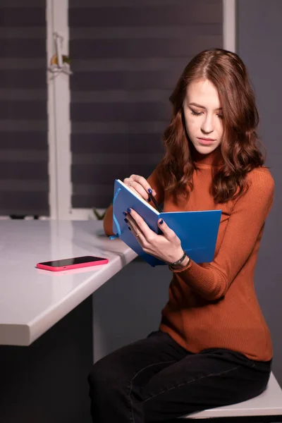 stock image charming brunette woman in brown golf with blue notebook and phone in pink case sitting at table in gray room. work, education, study. copy space