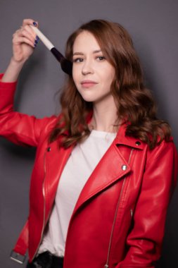 Kırmızı deri ceketli çekici esmer kadın makyaj fırçaları gri duvar arkasında izole edilmiş. Profesyonel makyöz. Boşluğu kopyala