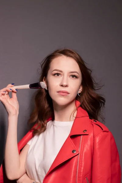 メイクブラシグレーの壁の背景に隔離された赤い革のジャケットの魅力的なブルネットの女性 プロの化粧師だ コピースペース — ストック写真