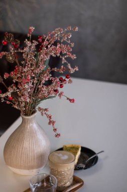 Süslü kırmızı dallı vazonun yanında bir fincan kahve ya da kapuçino. İç mekan, rahat. Boşluğu kopyala