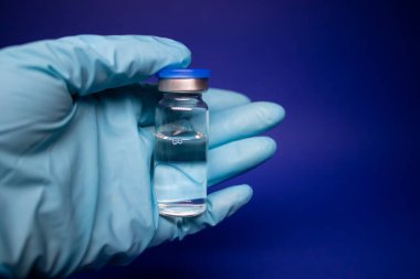 Koronavirüs aşısı COVID-19 'un gelişimi. Mavi tıbbi eldivende etiketi olmayan şişe şişesi. Mavi arka planda izole edilmiş. Tedavi. kopyalama alanı. Enfeksiyonların önlenmesi ve tedavisi için aşı