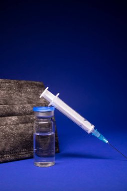 Siyah tıbbi maskenin yanında etiketsiz aşı şişesi şişesi ve enjeksiyonlu tıbbi şırınga. Mavi arka planda izole edilmiş. Tedavi. Koronavirüs aşısı COVID-19 'un gelişimi. Boşluğu kopyala