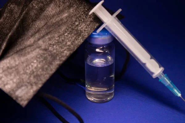 Vaccine bottle phial with no label near black medical mask and medical syringe with injection needle. isolated on blue background. cure. Development of coronavirus vaccine COVID-19. copy space