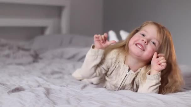 Feliz bonita niña rubia en vestido beige acostado en una cama en el dormitorio gris. rutina de la mañana, infancia, niño, hija. Imágenes de FullHD — Vídeo de stock
