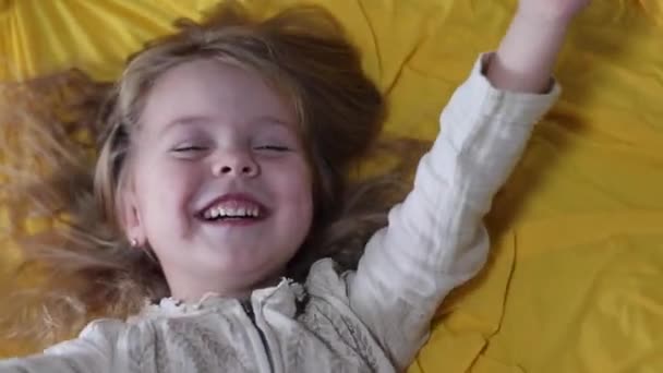 Glad liten blond flicka i pyjamas skrattar, liggande på gult silke textil. Morgonrutin, barndom, småbarn, dotter. FullHD-bilder — Stockvideo
