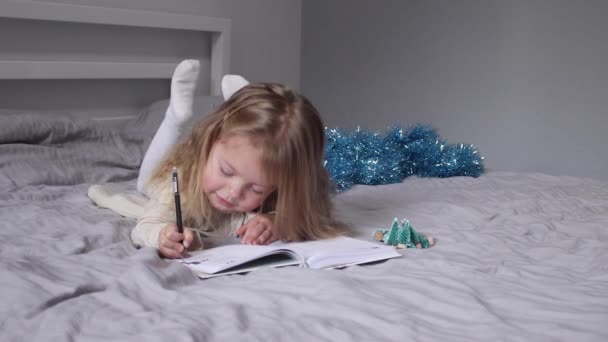 Очаровательная маленькая девочка пишет письмо Санта Клаусу на кровати в серой спальне. Новый год, детство, Рождество. FullHD — стоковое видео