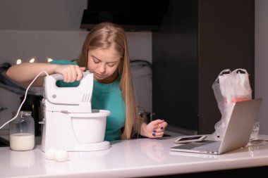 Modern mutfakta bilgisayar başında yemek pişiren sarışın bir kadın video blogcu, online öğrenme ya da uzaktan pişirme kursu konsepti. Canlı yayındayız. Karıştırıcıyı ve malzemeleri yakınında tut