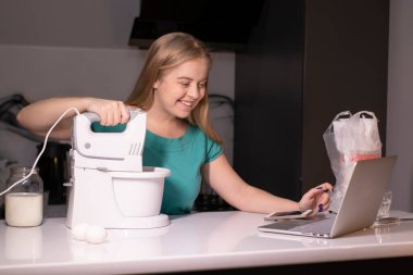 Modern mutfakta bilgisayar başında yemek pişiren sarışın bir kadın video blogcu, online öğrenme ya da uzaktan pişirme kursu konsepti. Karıştırıcı ve malzemelerin yanında dur. canlı yayın videosu