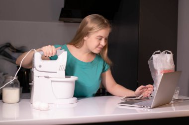 Modern mutfakta yemek pişirirken dizüstü bilgisayardan yemek tarifi izleyen genç sarışın kadın. Online aşçılık kursu. karıştırıcıyı ve farklı malzemeleri yakınlarda tut