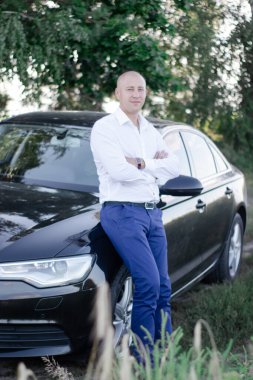 Lüks bir arabanın yanında takım elbiseli kel yakışıklı bir adam. Otomotiv işadamı