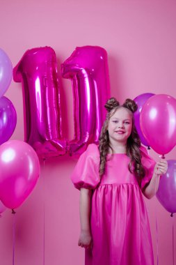 Pembe makyajlı ve pembe deri elbiseli tatlı esmer kız. Onbir numara şeklinde renkli balonların yanında. Pembe arka planda. Eğlence, parti, doğum günü kutlaması.