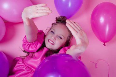 Pembe makyajlı ve pembe deri elbiseli çekici esmer kız modeli pembe arka planda renkli balonların yanında. Eğlence, parti, doğum günü konsepti.