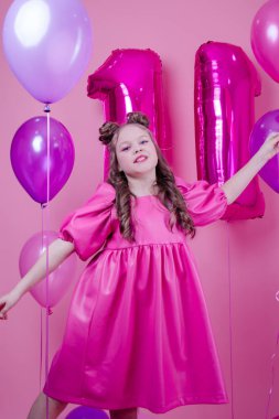 Pembe makyajlı ve pembe deri elbiseli çekici esmer kız modeli pembe arka planda renkli balonların yanında. Eğlence, parti, doğum günü konsepti.