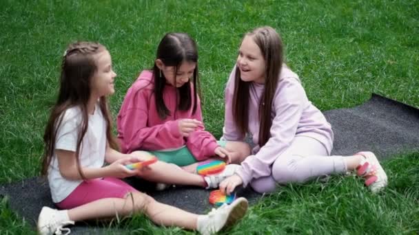 3 adolescentes en vêtements colorés jouent avec un jouet sensoriel tendance pop it outdor. bulles de silicone souple arc-en-ciel. soulagement de l'anxiété, antistress, concept de santé mentale. Jeu anti stress popit. au ralenti — Video