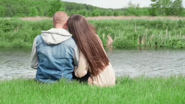 Dulce pareja sentada junto al río de agua sobre hierba verde. atractiva mujer morena y hombre calvo al aire libre. amor verdadero, familia feliz, romance, matrimonio exitoso. cámara lenta — Vídeo de stock