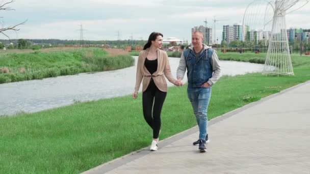 Härliga glada par promenader i parken vid floden håller hand. Brunett kvinna och skallig man. fru och man. lyckat äktenskap, sann kärlek. romantisk promenad på en dejt. slow motion — Stockvideo