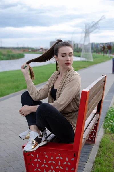 Ελκυστική μελαχρινή γυναίκα κάθεται σε ένα παγκάκι στο πάρκο. ρούχα μόδας, κομψή εμφάνιση — Φωτογραφία Αρχείου