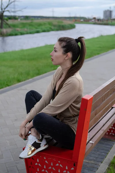 공원 벤치에 앉아 있는 매력적 인 갈색 머리 여인. 유행하는 의상, 유행하는 외모 — 스톡 사진